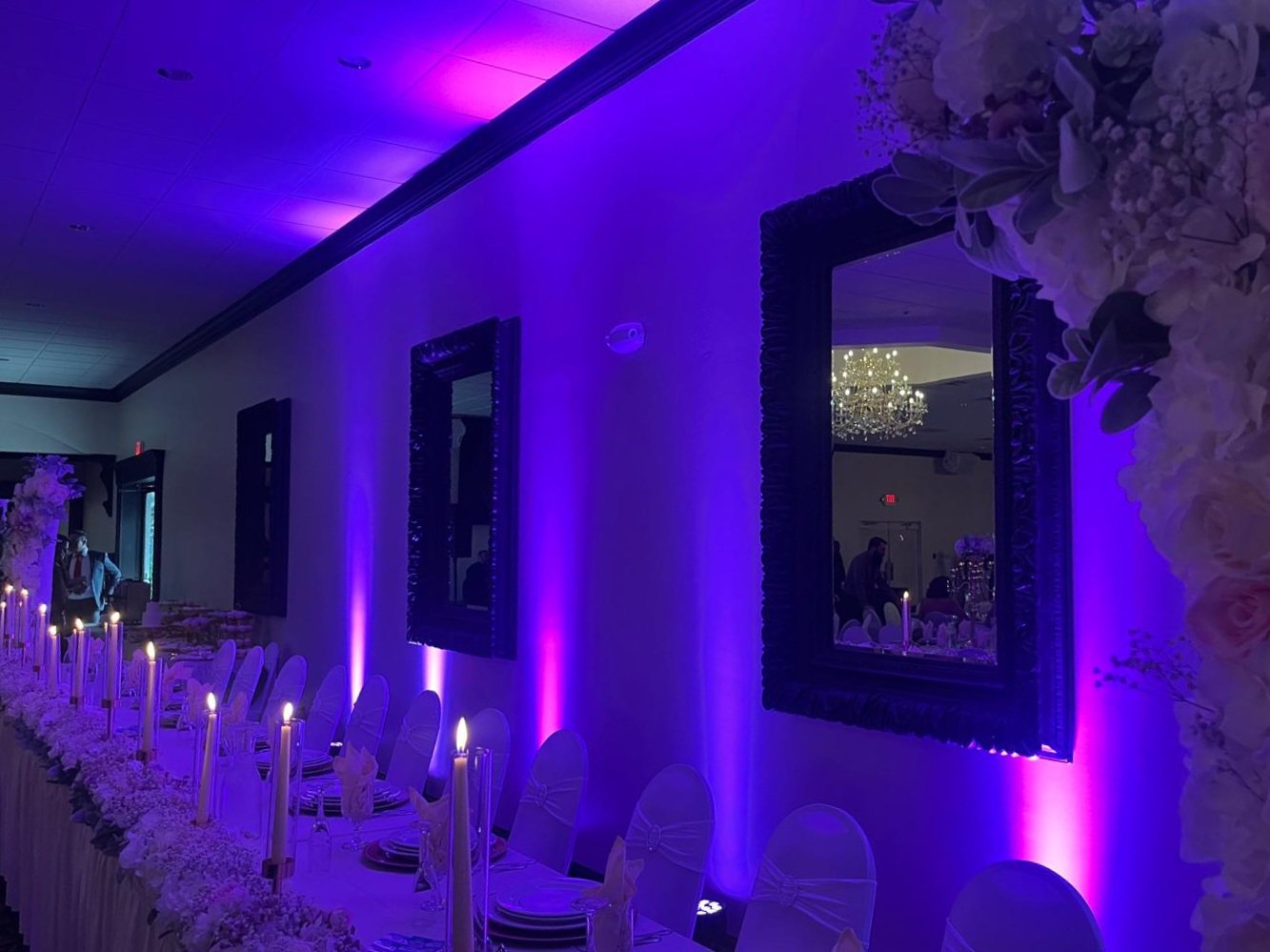 Decorated banquet room with purple lighting, a long table with white chairs, floral centerpieces, and mirrors on the wall reflecting a chandelier and guests.