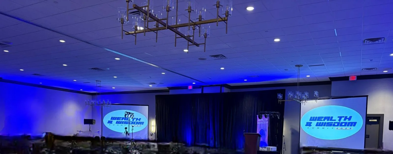 Conference room set up with two large screens displaying a logo for the 'Wealth & Wisdom Summit 2023.' The room has blue lighting, chandeliers, and a stage area with a podium.
