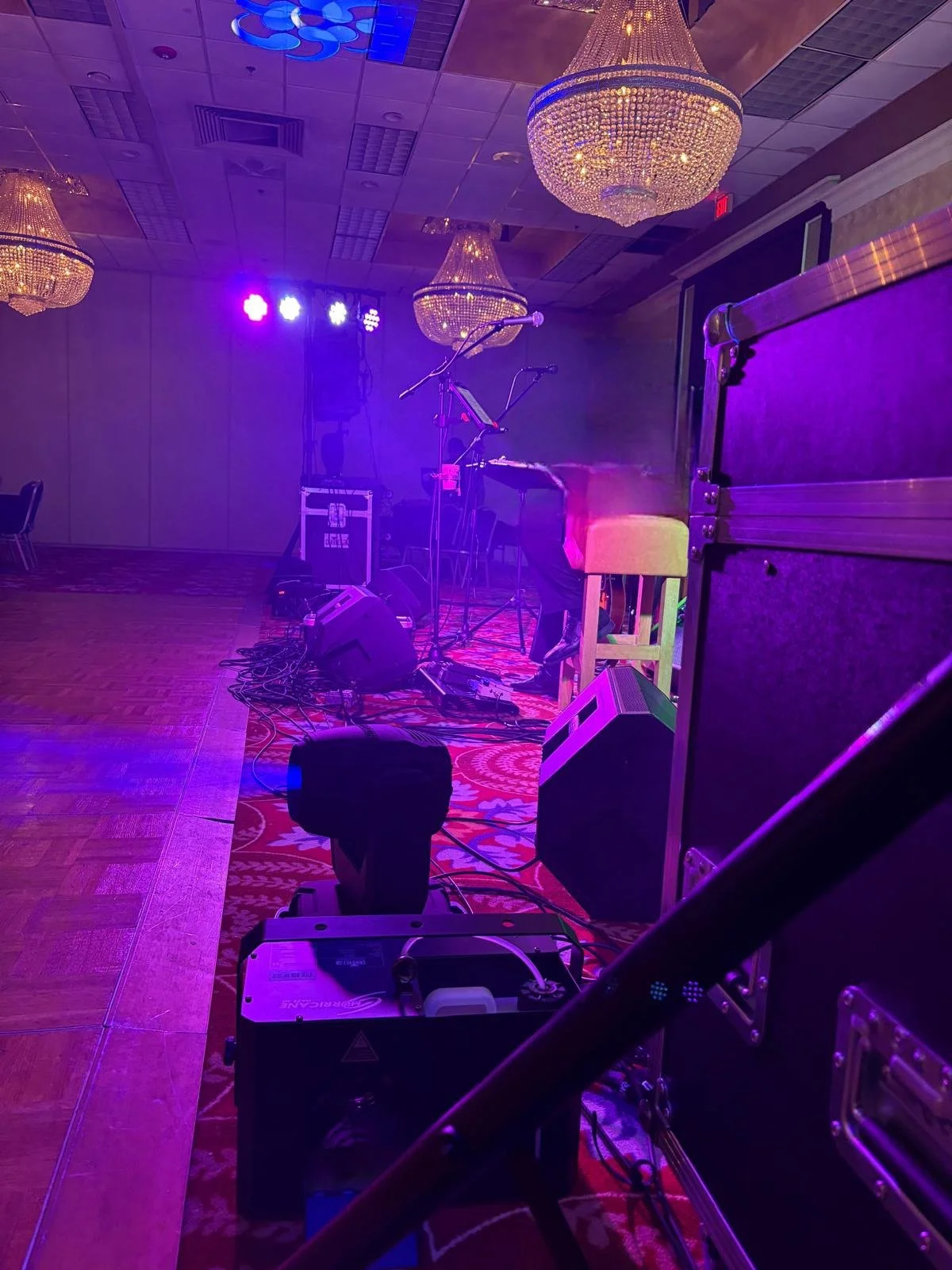 Stage setup with lighting, musical instruments, microphone stands, and speaker cabinets in a ballroom with chandeliers.