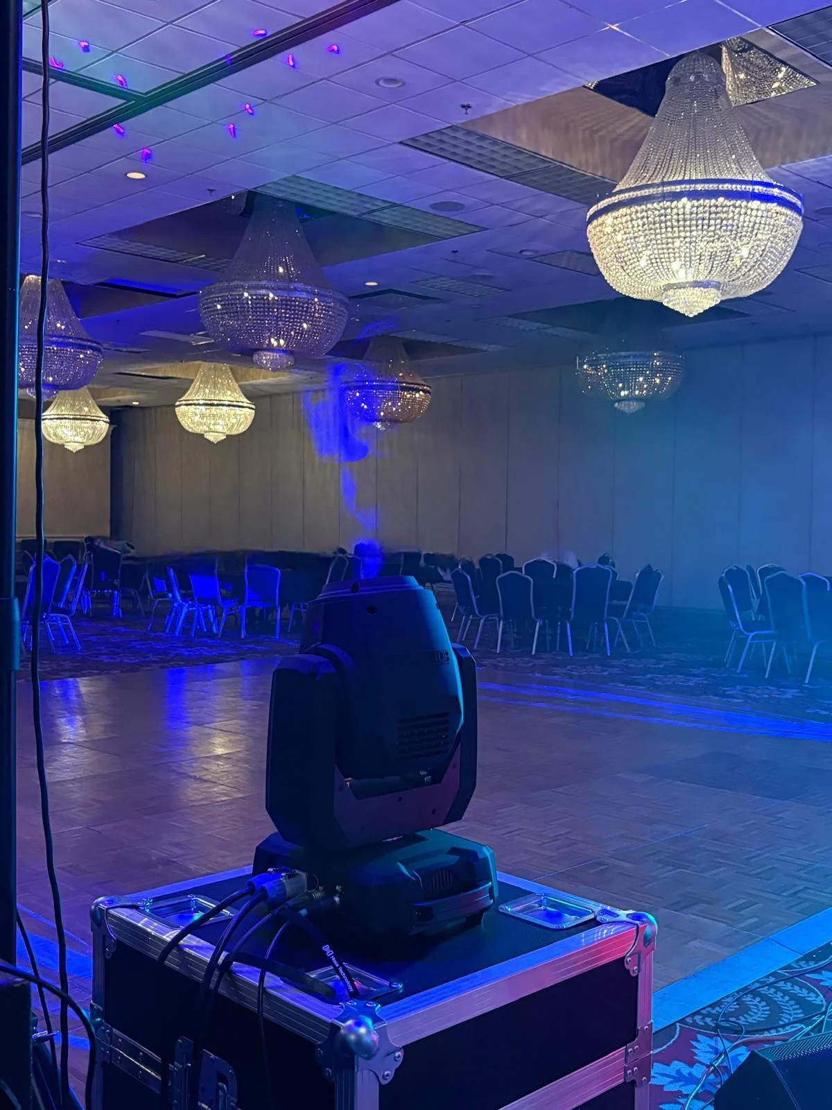 Empty event hall with several large chandeliers hanging from the ceiling, a neutral-colored wall, and chairs arranged in rows. There is a stage lighting fixture in the foreground.