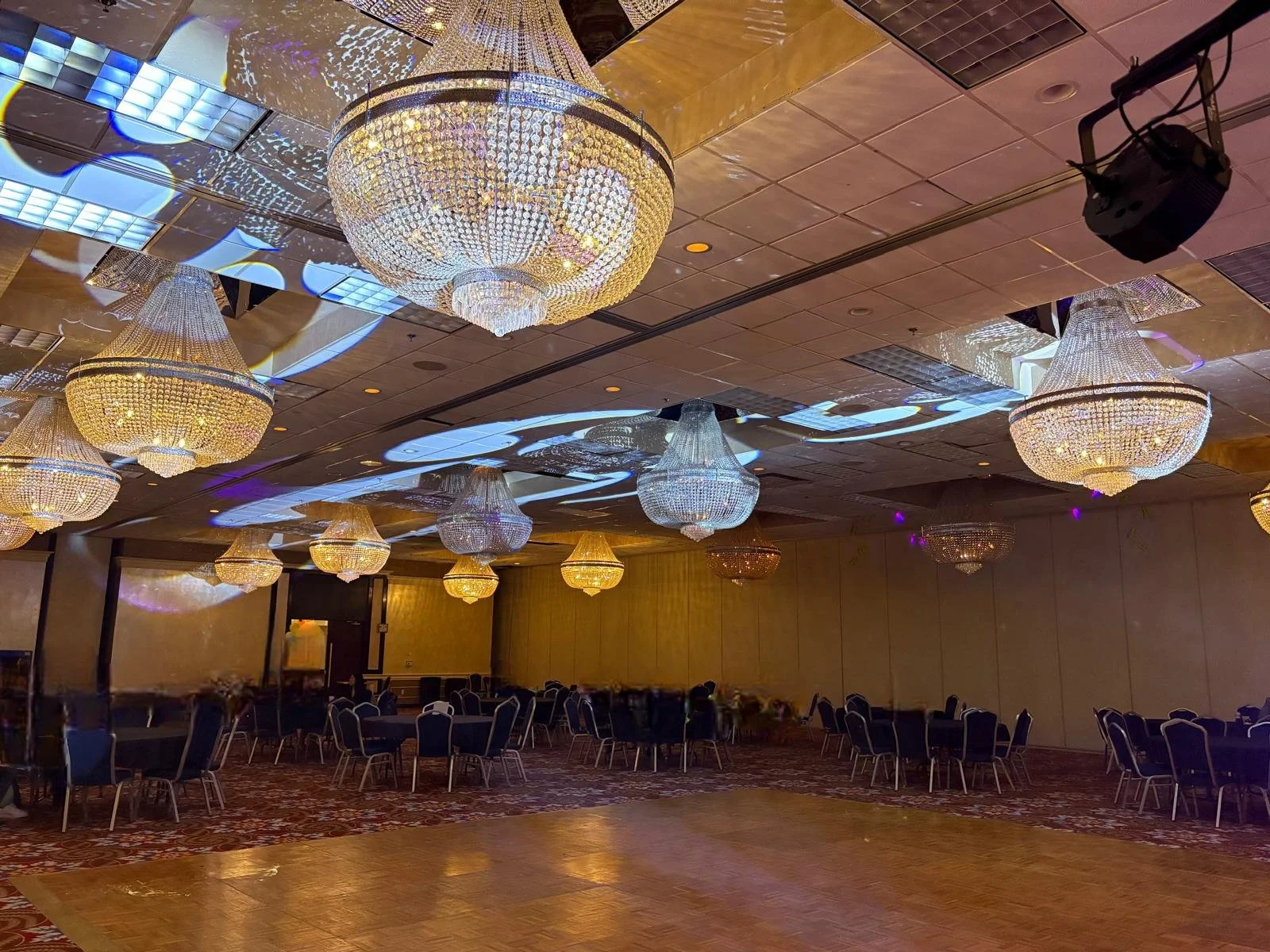 A large banquet hall decorated with multiple chandeliers hanging from the ceiling, reflecting colorful lights onto the ceiling and walls. The hall has round tables and chairs arranged for an event.