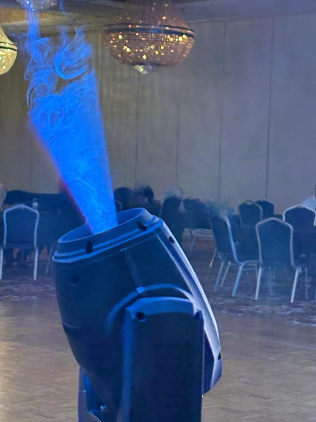 A fog machine emitting blue smoke in an indoor event space with chairs and chandeliers.