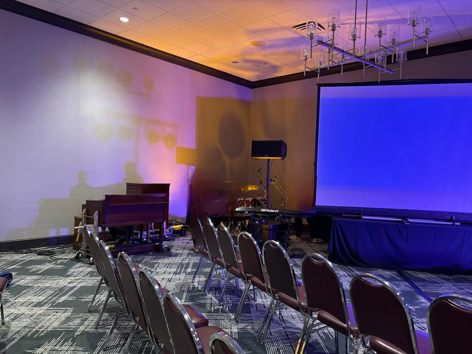 An empty conference room with chairs facing a large blue projection screen, stage equipment, and lighting setup in the corner.