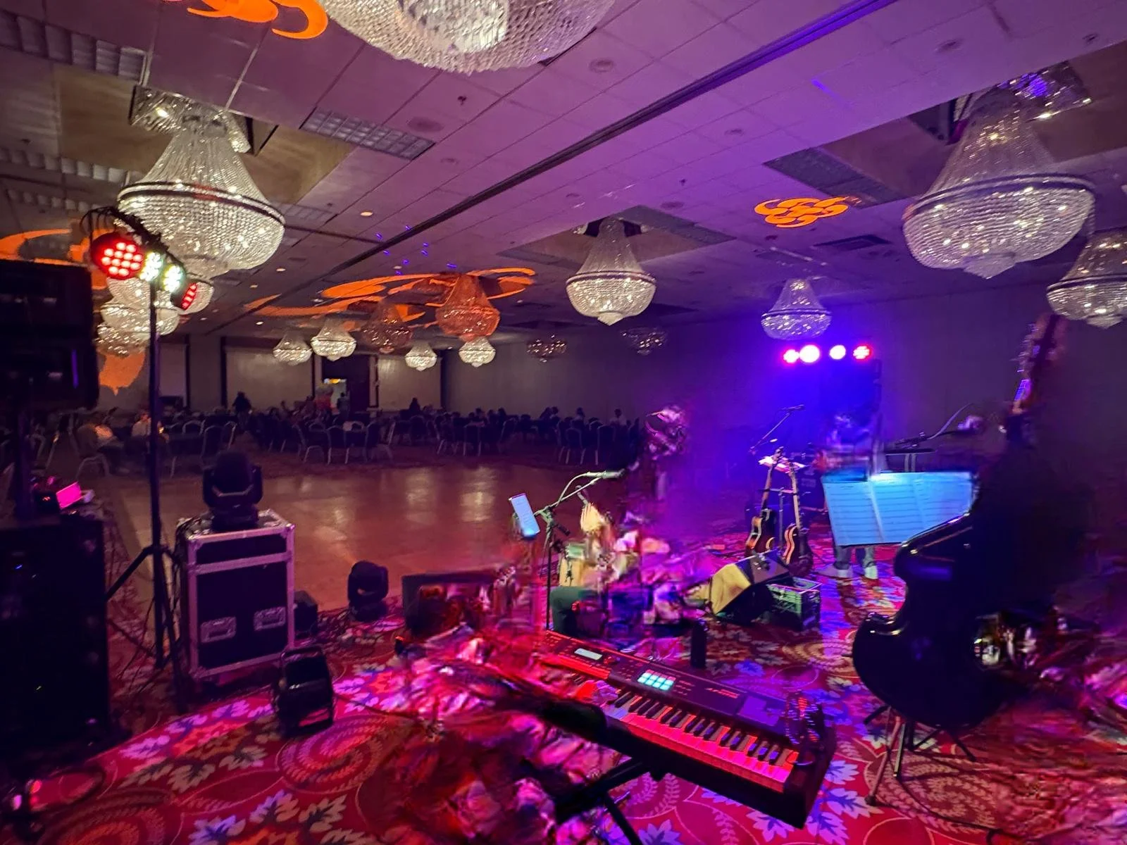 Empty event hall with chandeliers, stage setup with musical instruments, keyboard, guitars, drum set, and microphone, with colored lights and a purple glow, and a few spotlights.