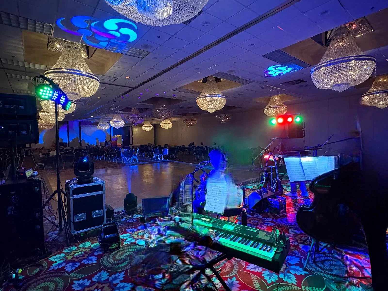 Empty banquet hall with chandeliers, stage with piano, microphone, sheet music, and colorful DJ lights, set up for a performance or event.