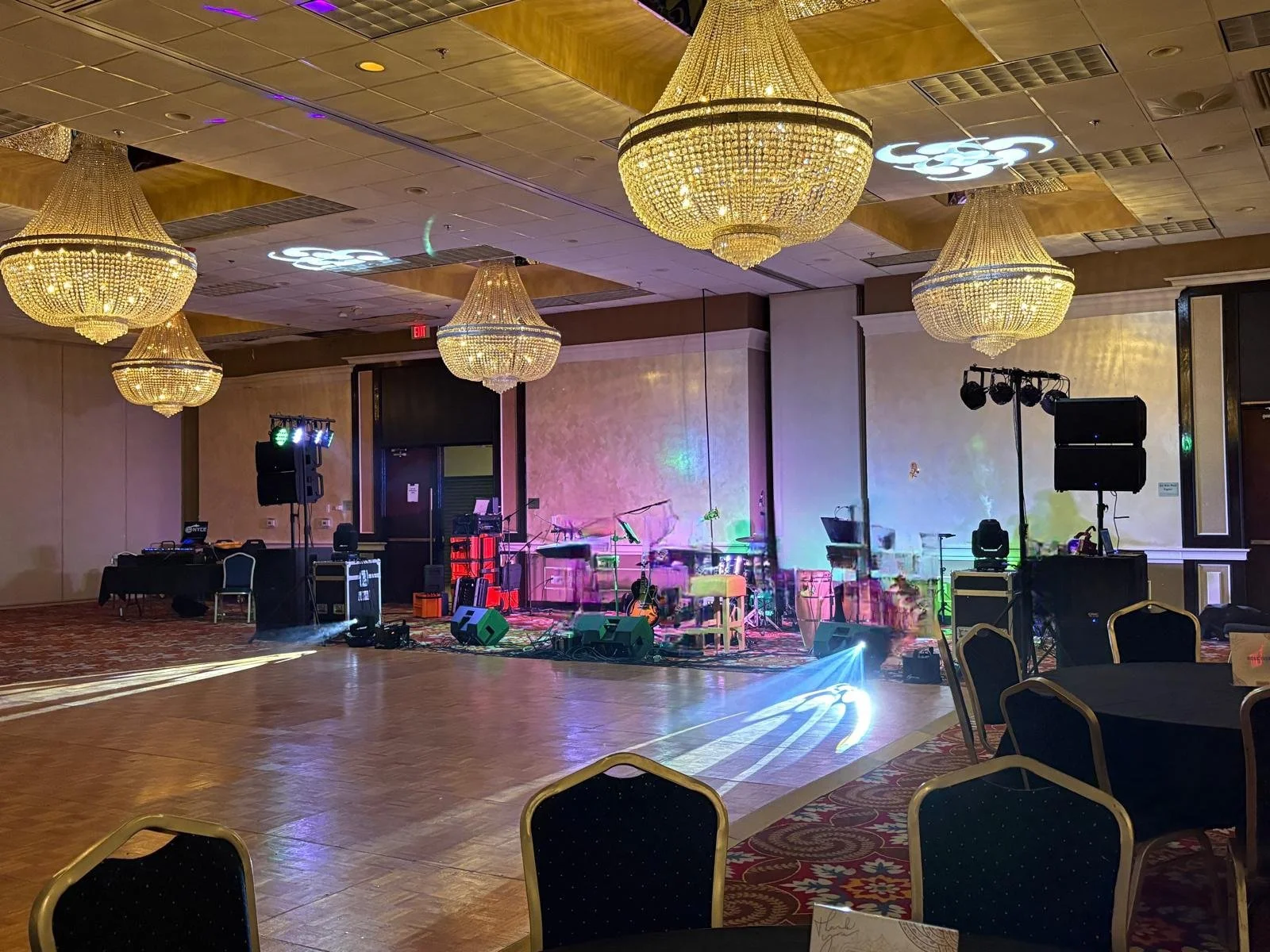 An elegant banquet hall with multiple large chandeliers, a stage with musical instruments including microphones, speakers, and lighting effects, and round tables with chairs set up for an event.
