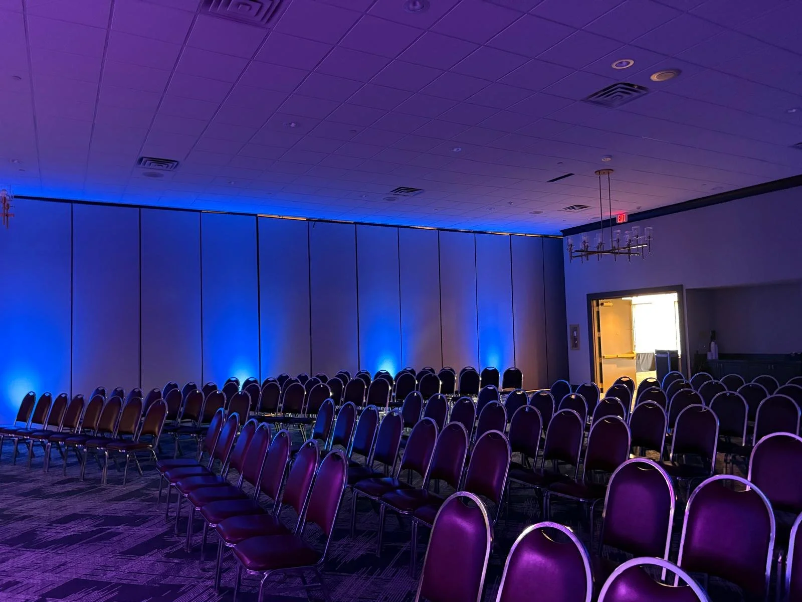 Empty conference room with rows of purple chairs facing a wall illuminated with blue lights, and a doorway leading to a brightly lit room.