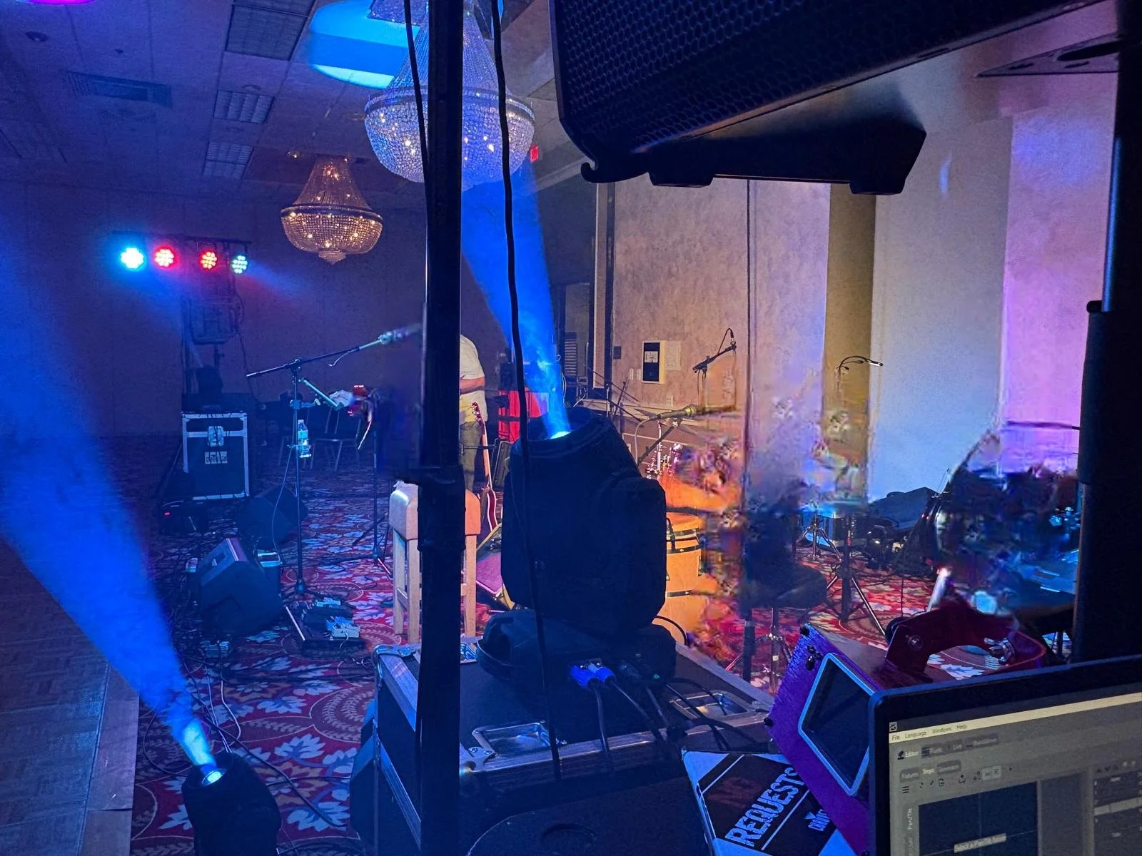 Stage setup with musical instruments, microphones, and colorful stage lighting, inside a banquet hall with chandeliers and decorative carpet.