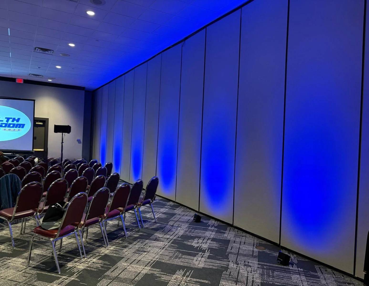 Empty conference room with rows of chairs, a screen displaying a logo, and blue lighting along the wall.