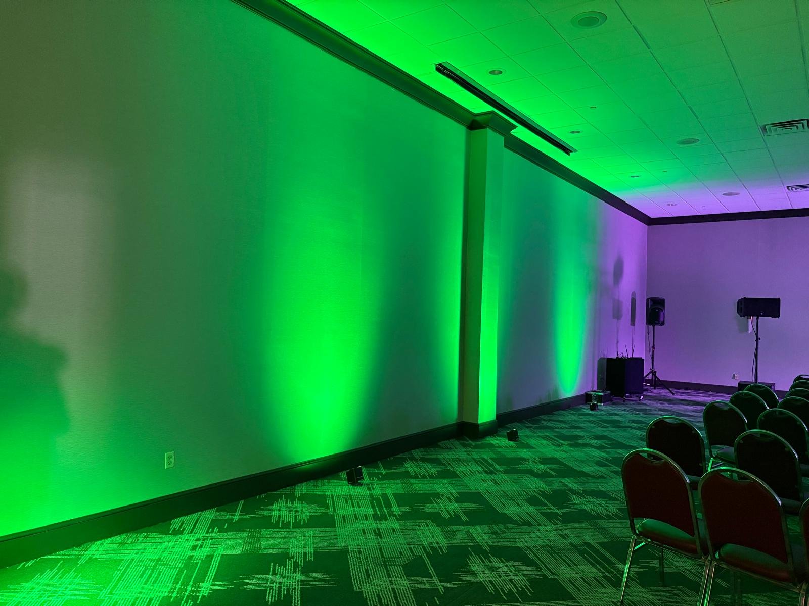 An empty conference room with green and purple spotlights on the walls, black chairs arranged in rows, and speakers on stands.