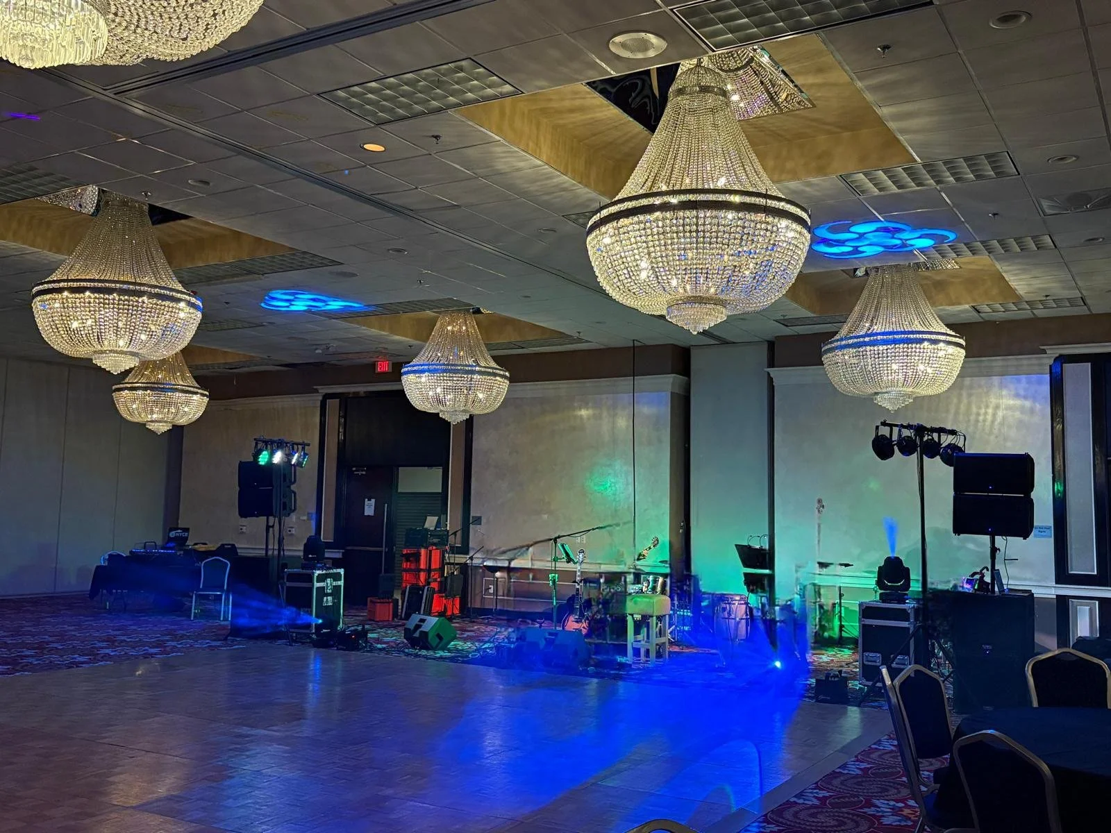 A banquet hall or event space with four large, ornate chandeliers hanging from the ceiling, and a stage with lighting and sound equipment set up for a performance or DJ. The room is dimly lit with blue and green stage lights.