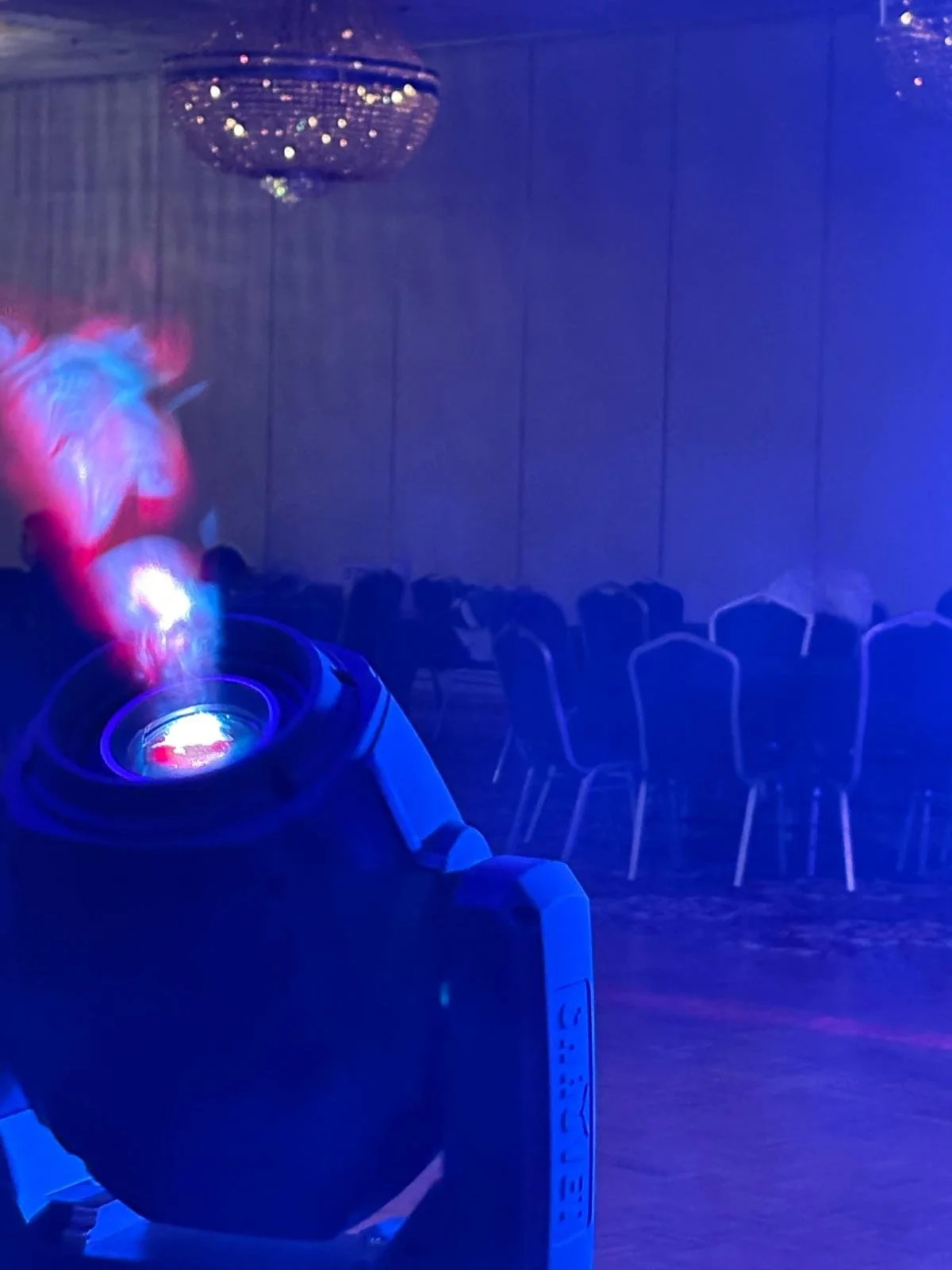 A disco ball hangs from the ceiling, and a fog machine emits colorful smoke in a dimly lit room with purple and blue lighting, empty chairs arranged in rows, likely set up for a party or event.