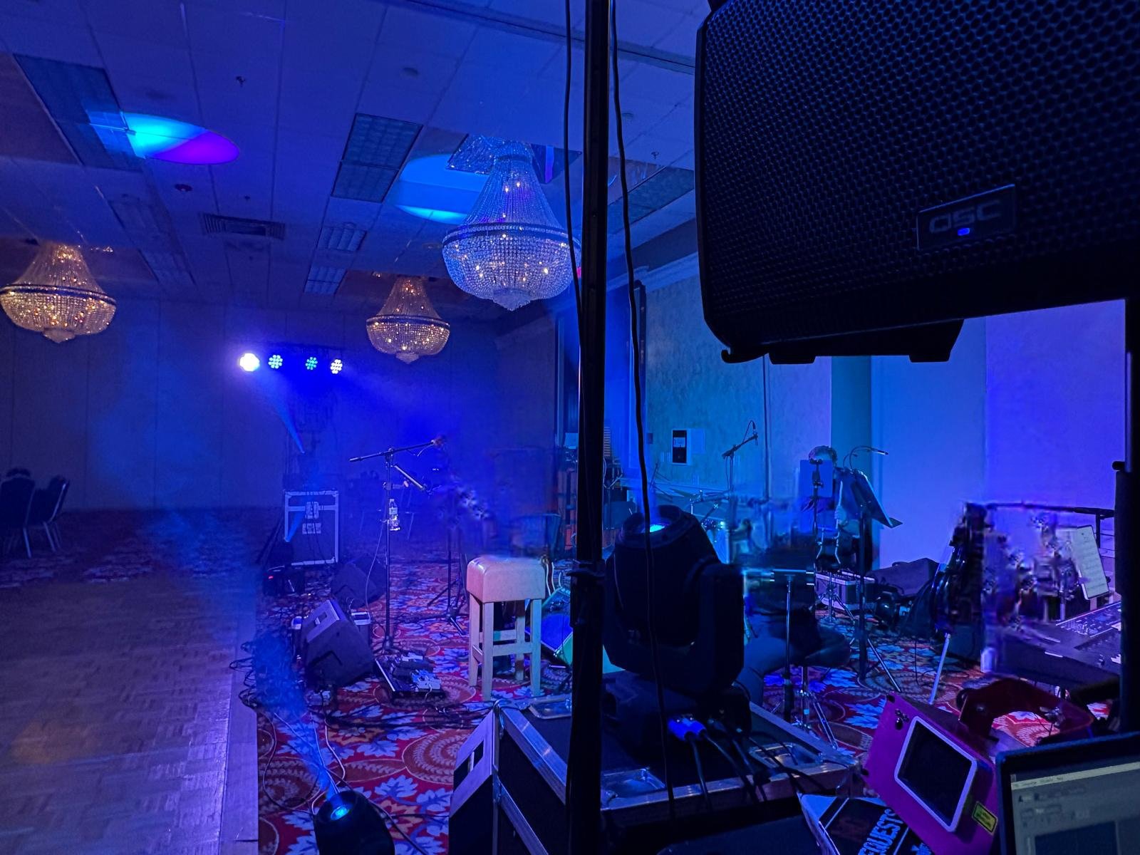 Empty event stage with musical instruments and equipment, illuminated by blue and purple lighting, with chandeliers hanging from the ceiling.