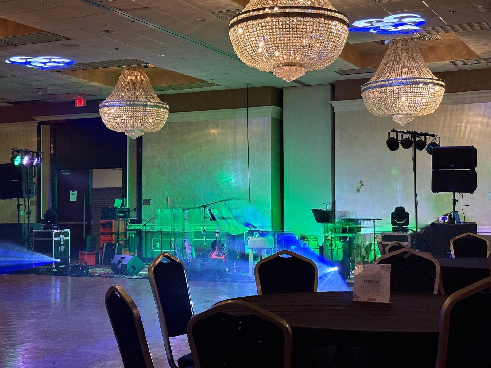 Empty banquet hall with multi-colored stage lighting, three large chandeliers hanging from the ceiling, and a reserved table in the foreground.