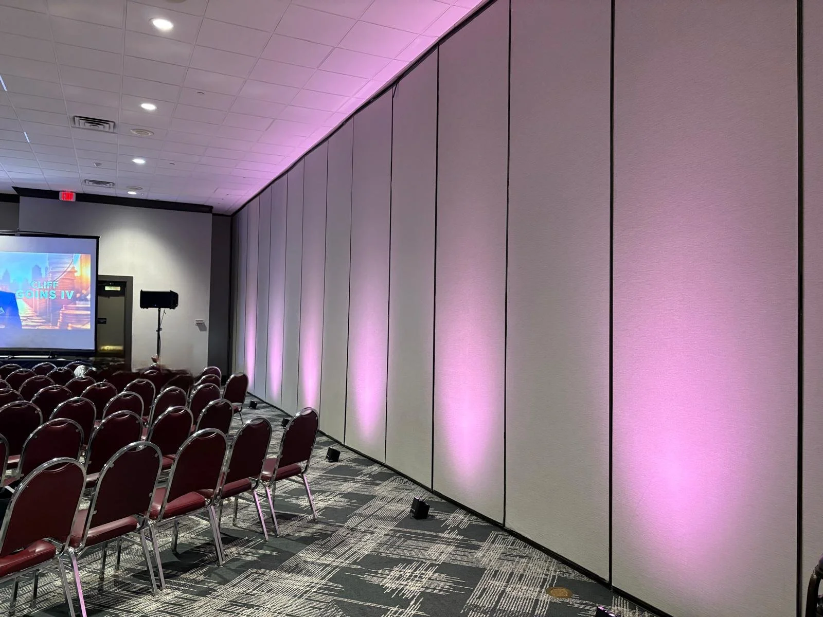 Empty conference room with rows of red chairs facing a large screen and a speaker, and soft pink uplights illuminating the beige wall near the ceiling.