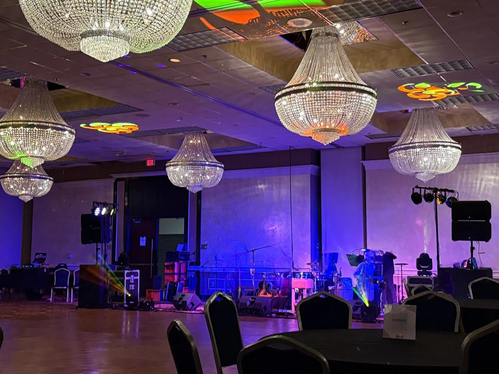 Empty banquet hall with large chandeliers, a stage with musical equipment, purple lighting, and some chairs and tables set up, possibly for a performance or event.