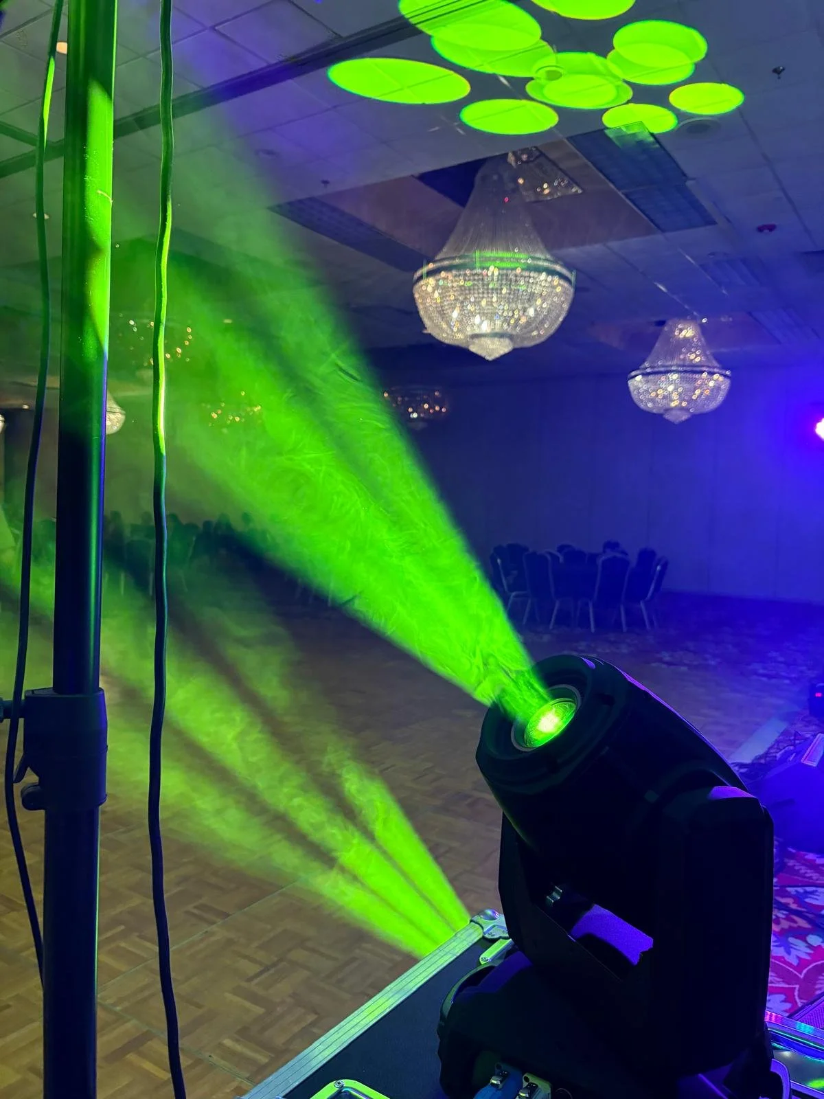 Stage lighting with a green spotlight projecting onto a dance floor, chandelier lights hanging from the ceiling, and empty chairs in the background of a ballroom or event space.