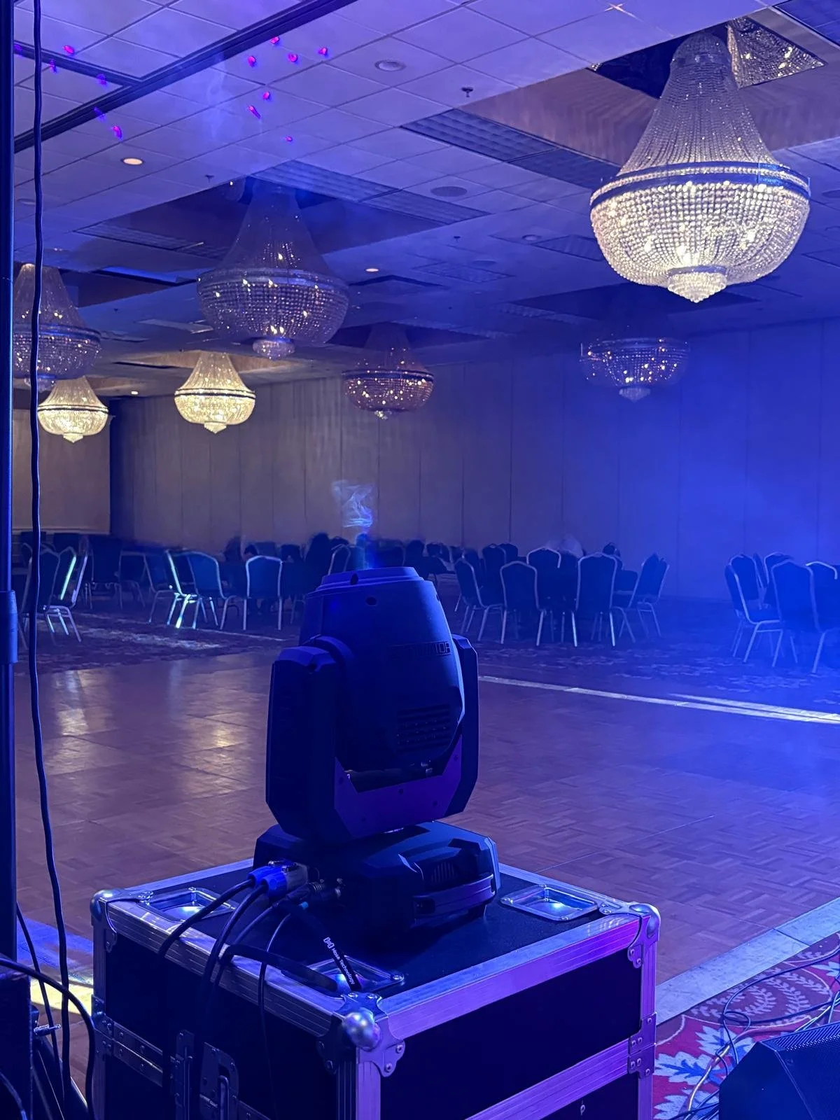 Empty banquet hall with large chandeliers, arranged chairs, and a fog machine on the floor, with a blue hue lighting.