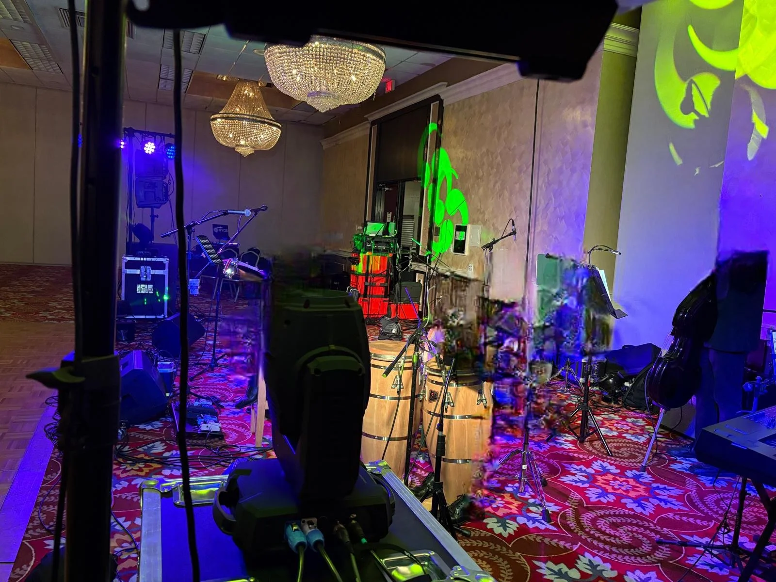 Stage setup at an event with musical instruments including drums, a keyboard, microphones, and amplifiers, illuminated with colorful stage lighting in a room with chandeliers and decorative carpeting.