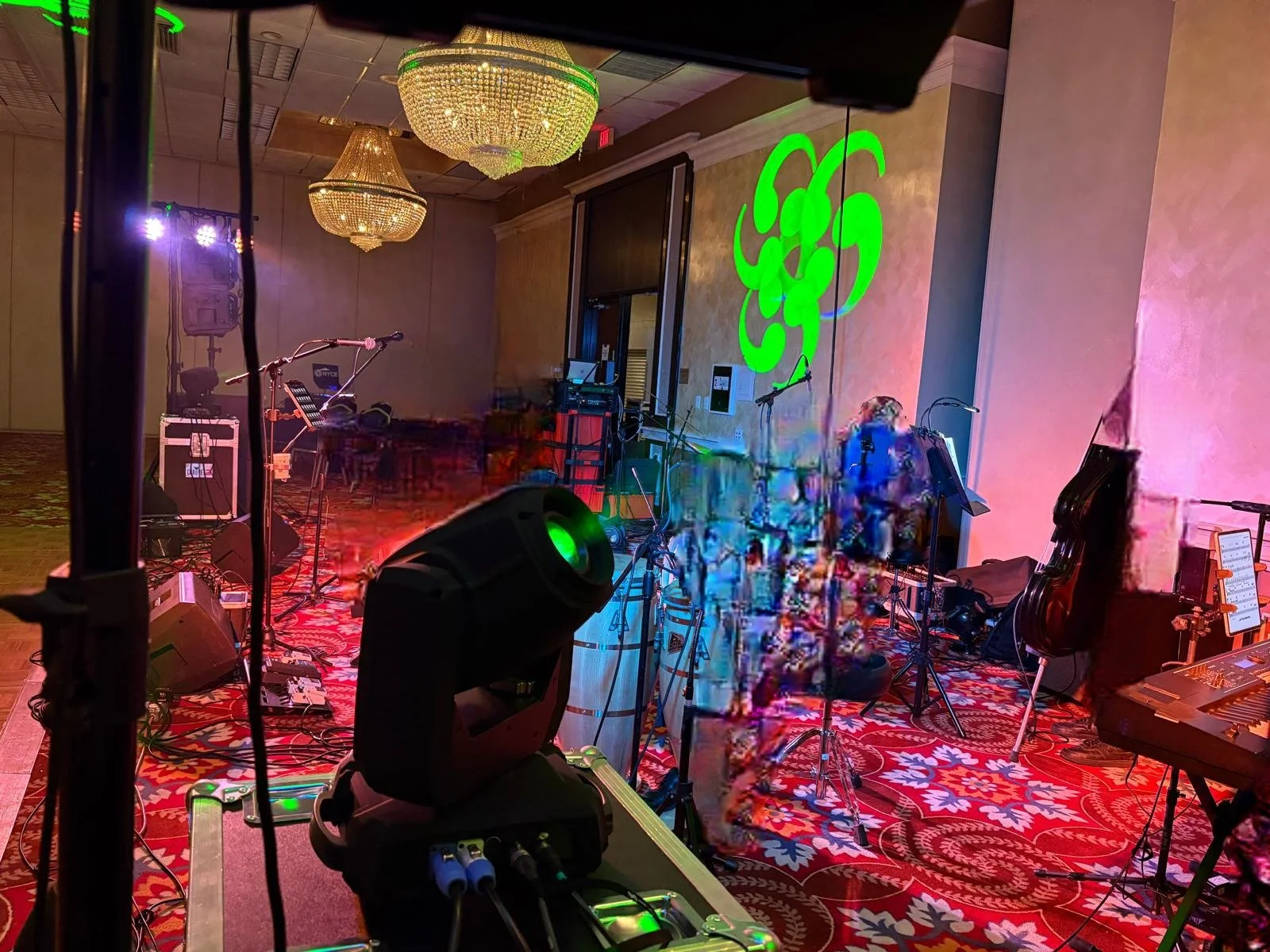 Empty stage set up for a musical performance with musical instruments, microphones, and stage lighting, in a ballroom with chandeliers and a patterned carpet.