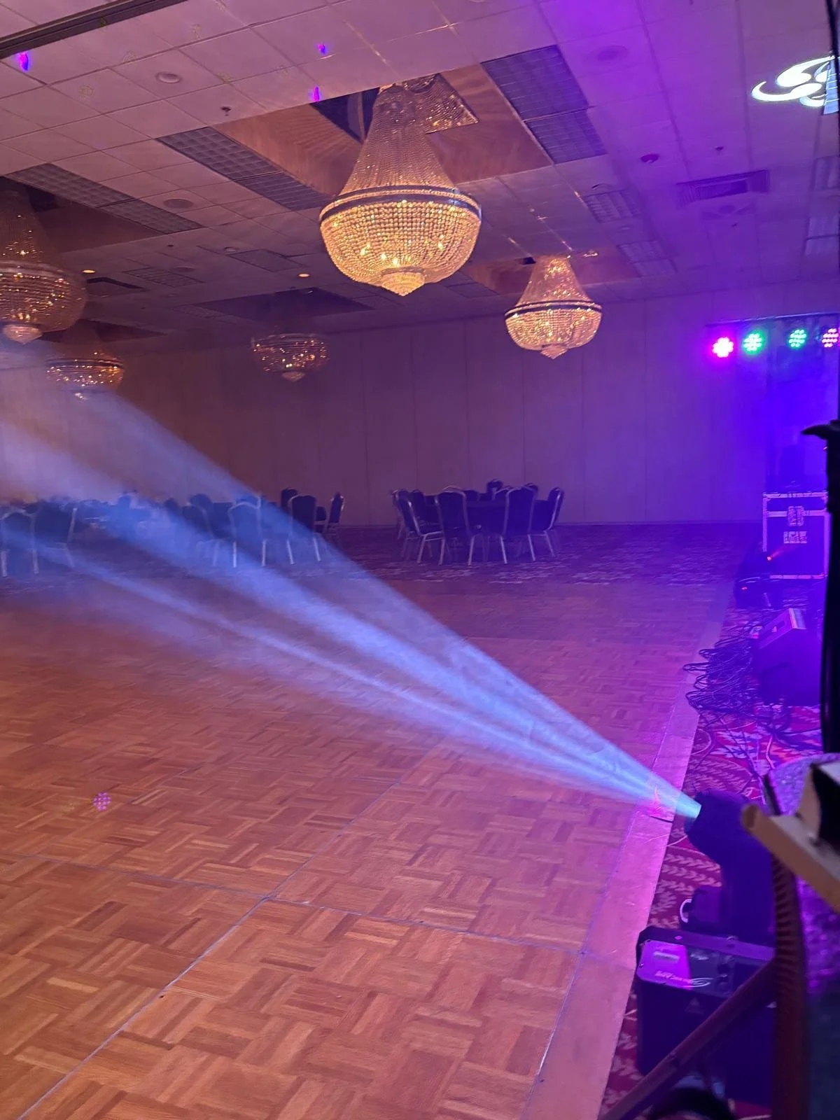 Empty banquet hall with chandeliers, empty tables and chairs, colorful stage lighting, and a fog machine on the dance floor.