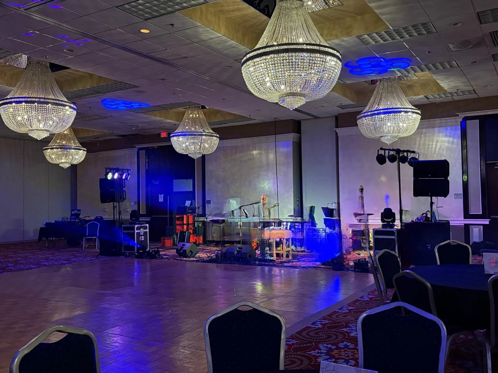 Empty banquet hall with large crystal chandeliers, stage with music equipment, blue lighting, and empty chairs and tables.