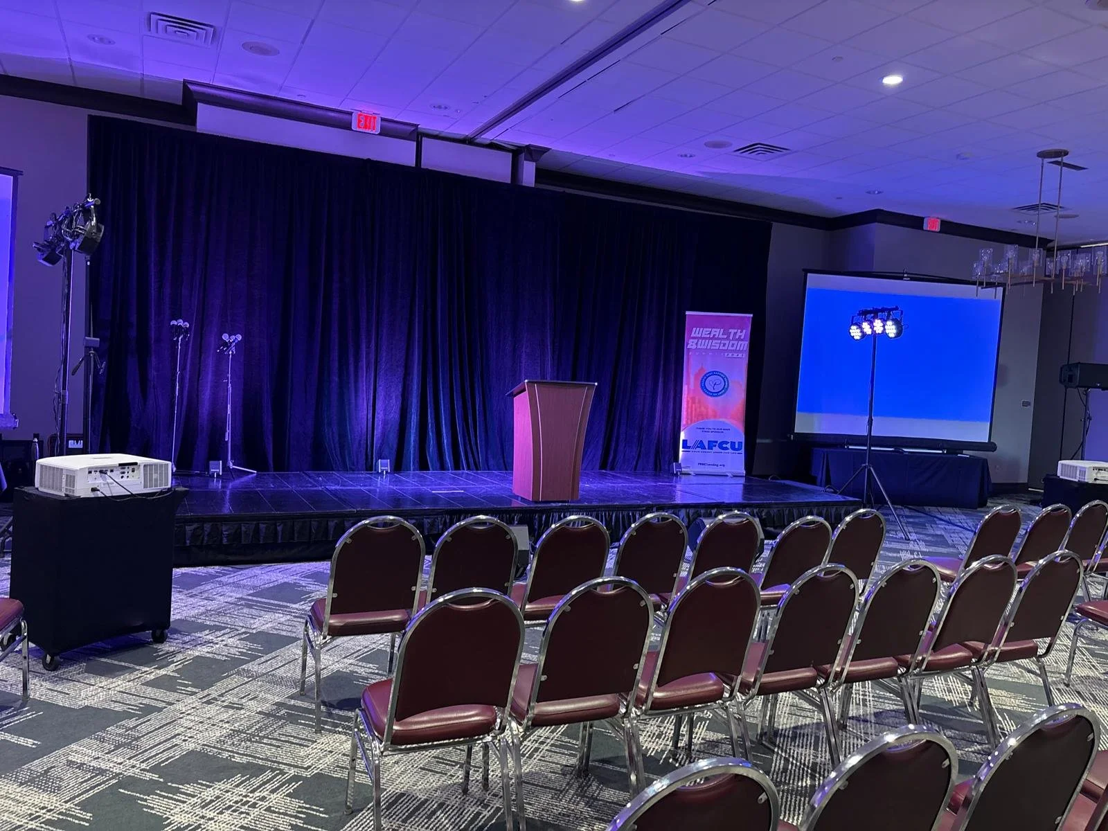 Empty stage with purple curtains, podium, projector, large screen, and chairs set up for an event in a conference room.