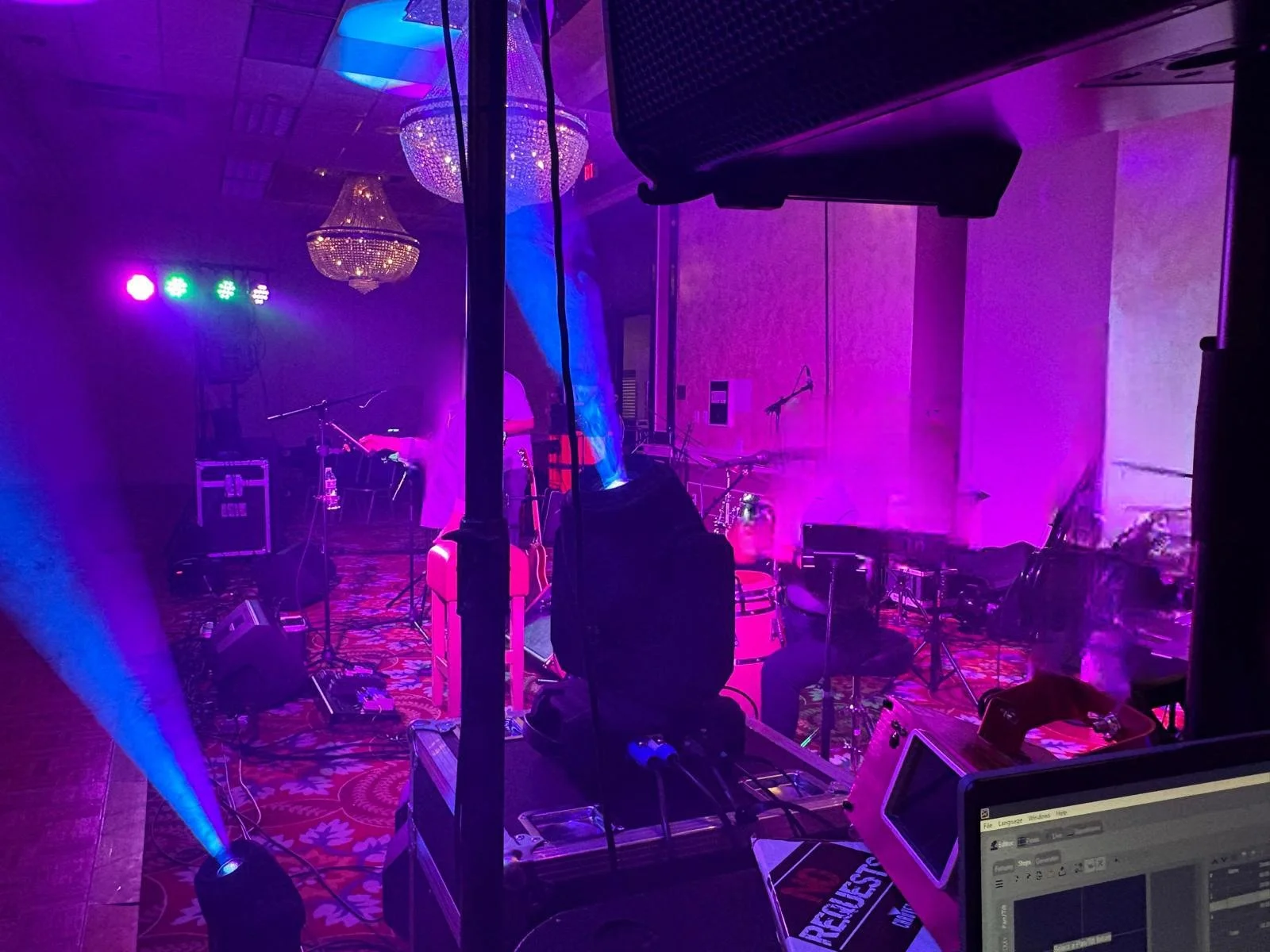 Stage with musical instruments, microphones, and lighting equipment set up in a room illuminated with purple, pink, and blue lights. Chandeliers hanging from the ceiling add elegance to the scene.