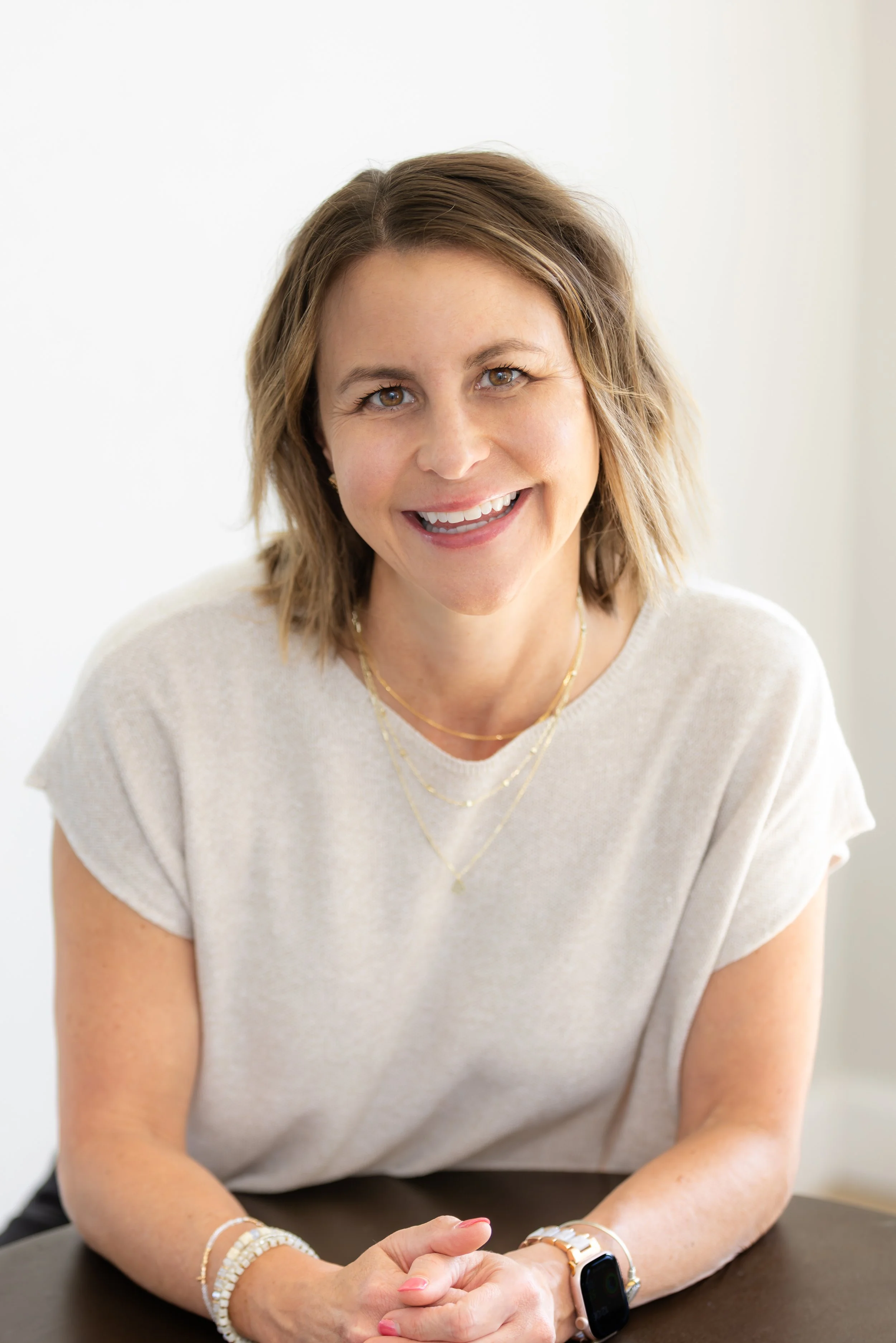 A woman with shoulder-length light brown hair smiling, wearing a cream-colored top, gold necklaces, bracelets, and an Apple Watch, sitting at a table.