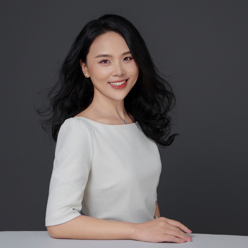 A woman with long black hair, smiling, wearing a white top, posed against a plain gray background.