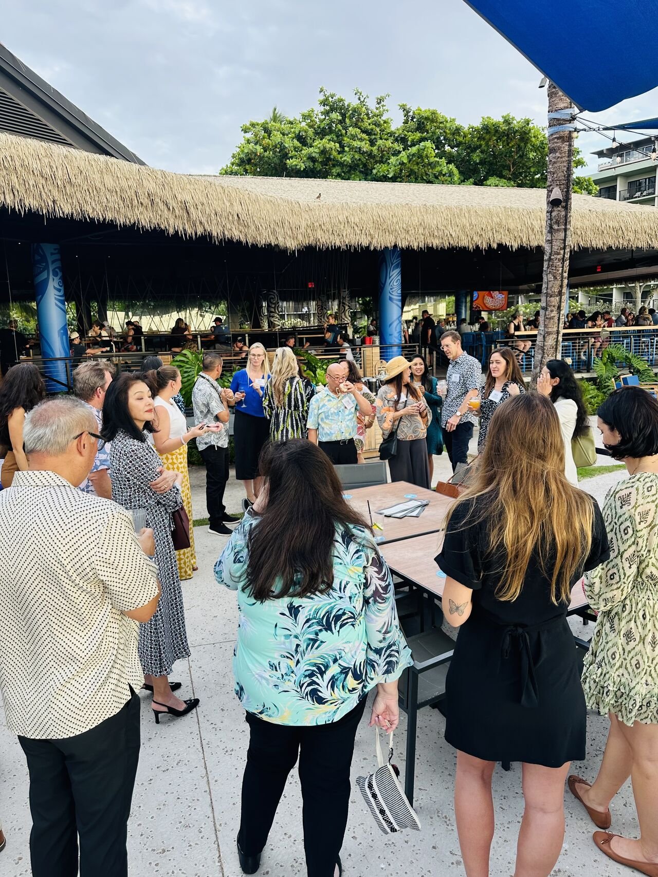 A group of people gathered outdoors near a bar at a tropical-style venue, some are holding drinks, with a thatched roof overhead and trees in the background.