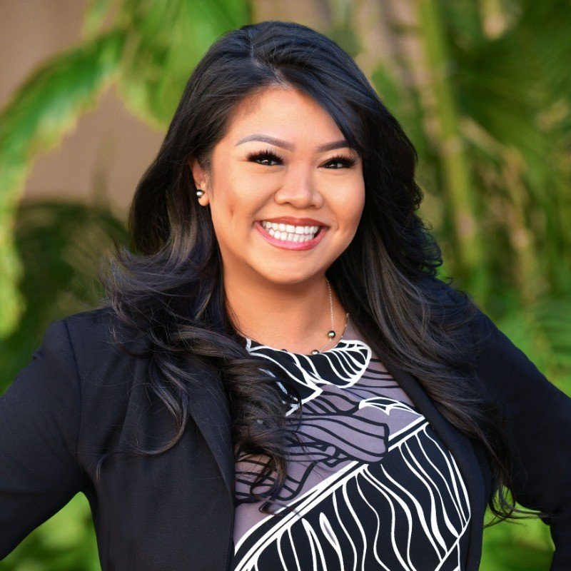 A woman with long dark hair smiling outdoors with green foliage in the background, wearing a black blazer and a patterned blouse.