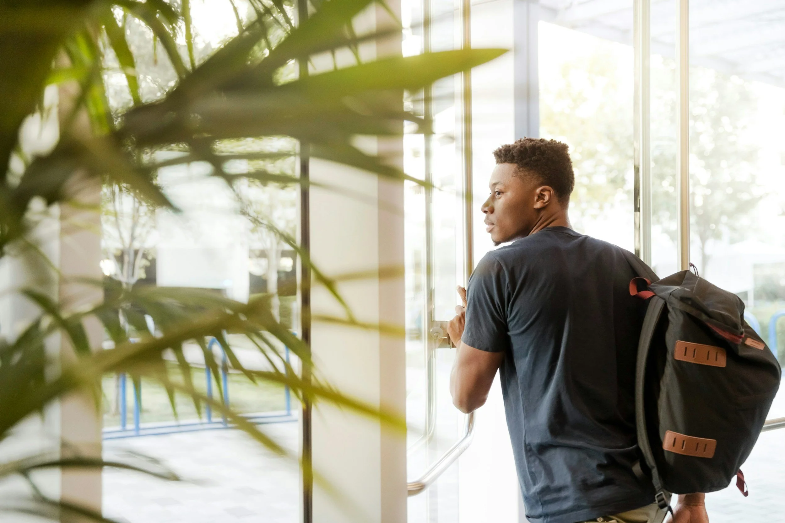 A young man with a backpack looking out of a window in a bright indoor space.