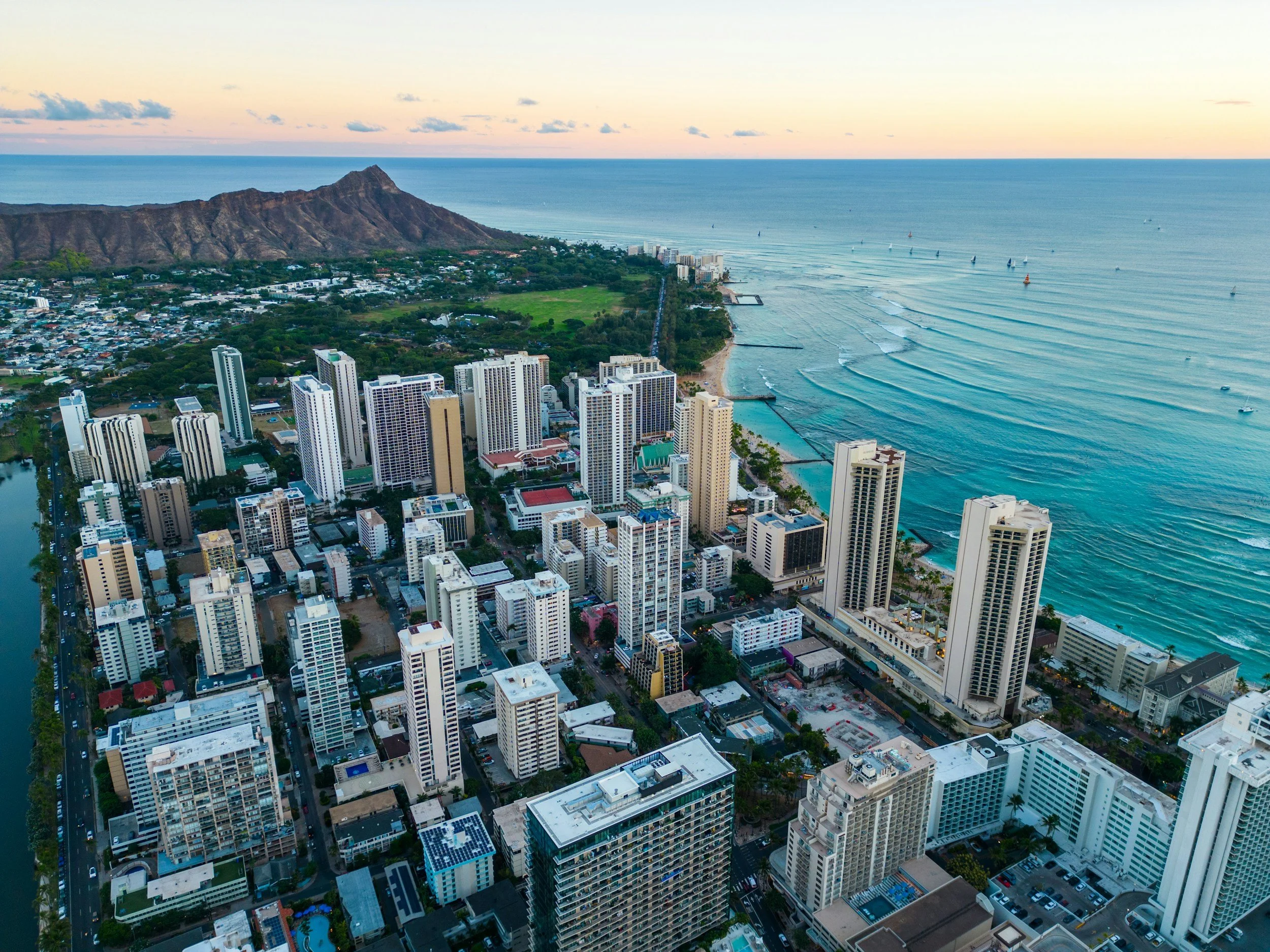 Aerial view of Waikiki cityscape with high-rise buildings along the shoreline and the Pacific Ocean, with Diamond Head crater in the background during sunset.