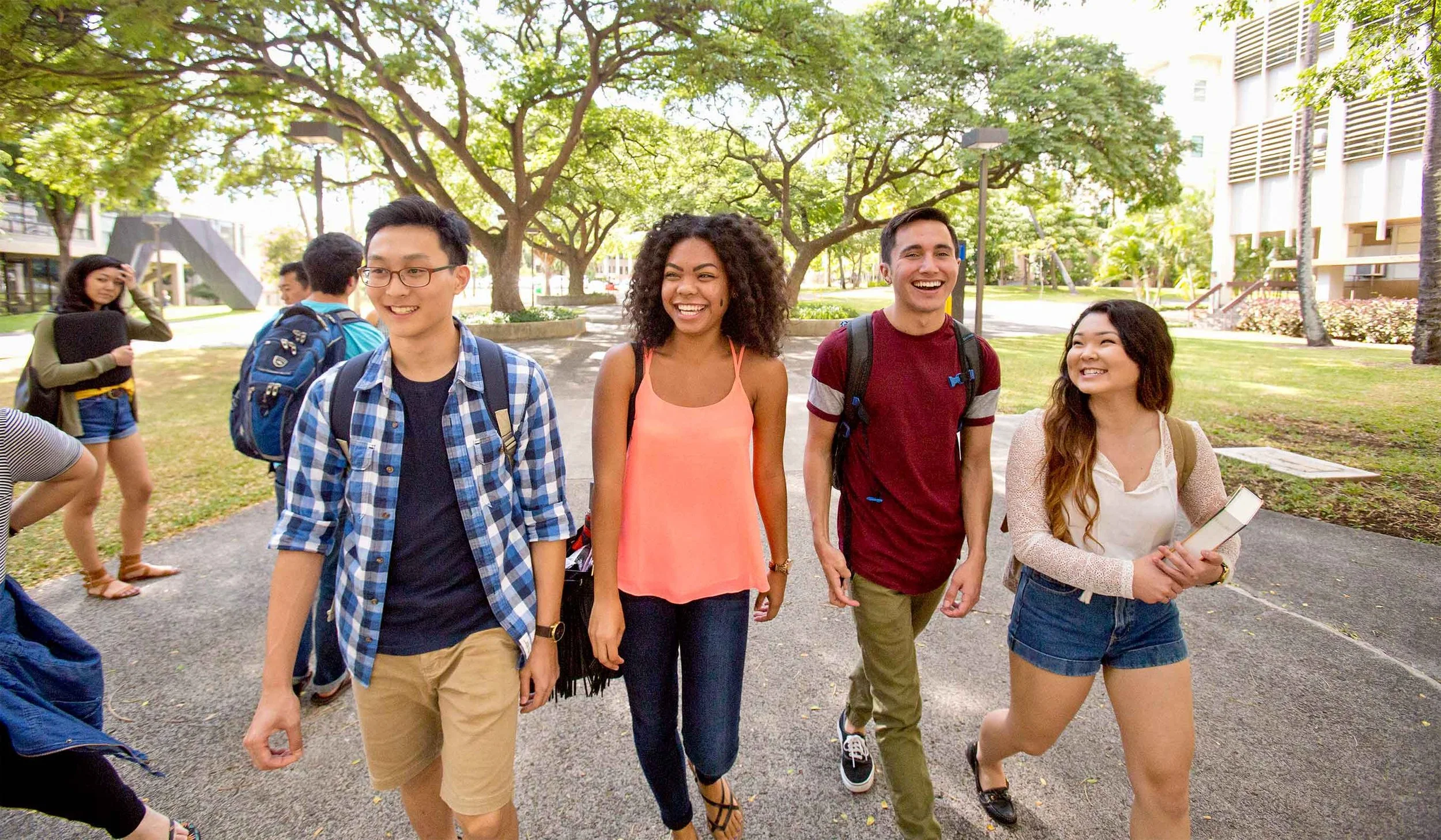 Four diverse students walk and chat outdoors on a sunny college campus with green trees and modern buildings in the background.