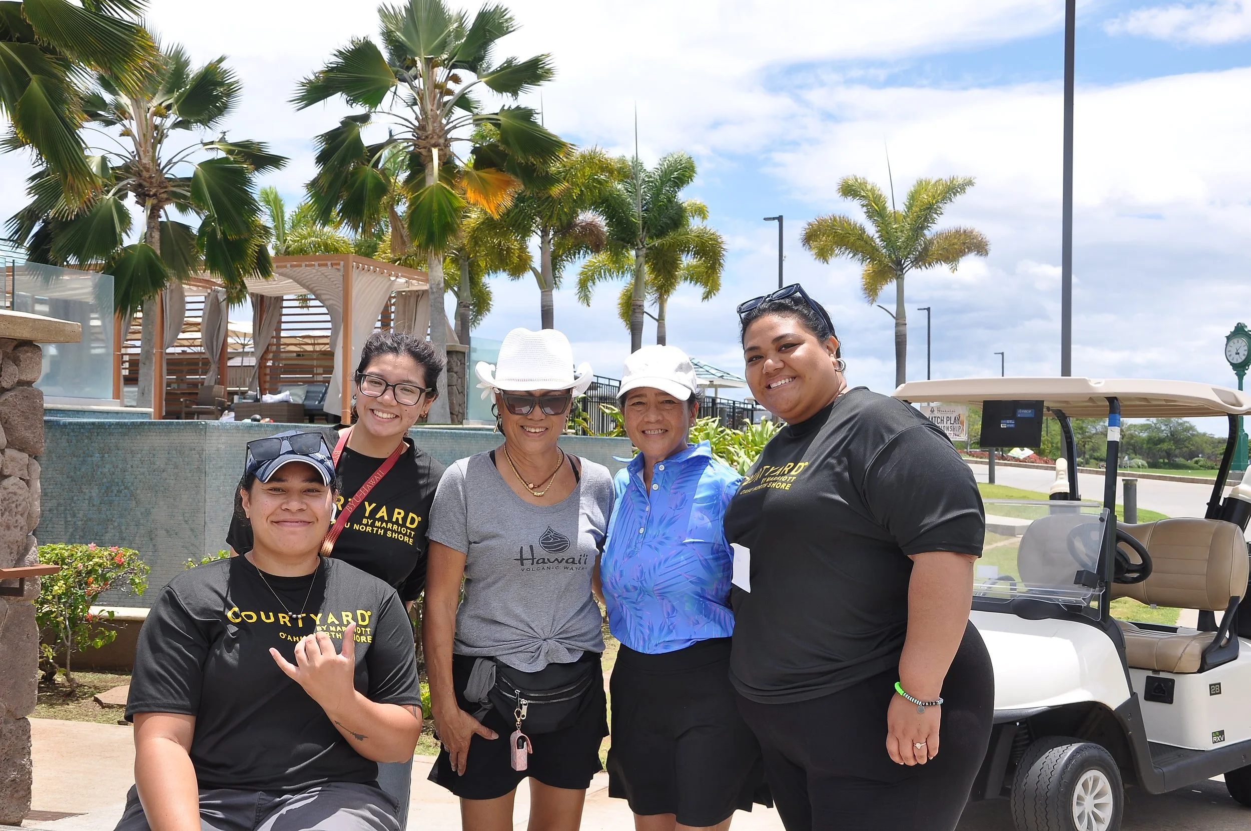 Five women standing outdoors near a golf cart, smiling and posing for the photo, with palm trees and a blue sky in the background.
