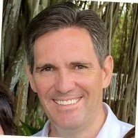 A smiling man with short gray hair, wearing a white shirt, outdoors near trees.