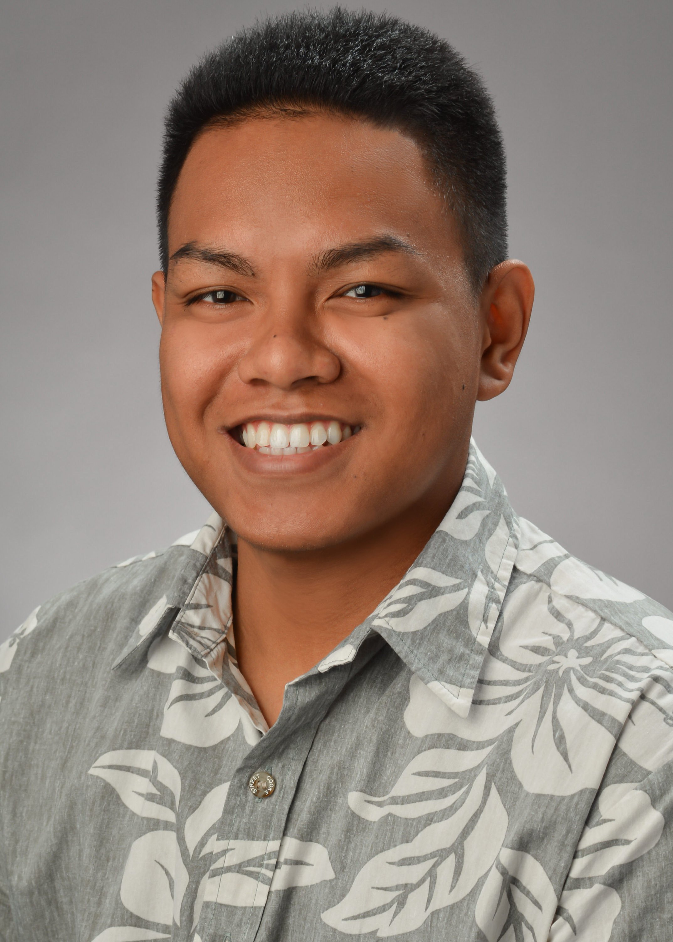 Smiling young man with short dark hair, wearing a gray Hawaiian shirt with white floral patterns, against a plain gray background.