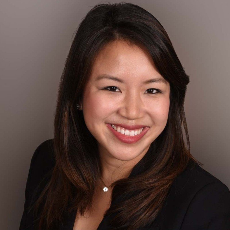 Headshot of a smiling Asian woman with shoulder-length dark hair, wearing a dark blazer and a necklace, against a neutral background.
