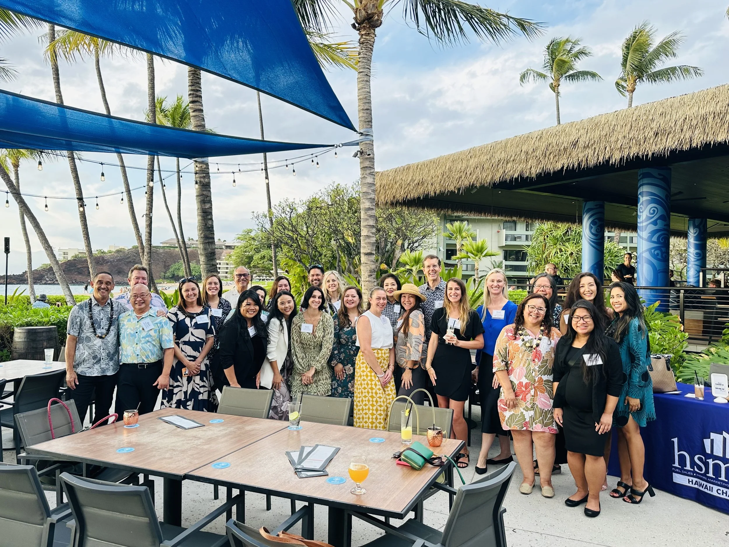 Group of people gathered outdoors at a tropical venue with palm trees, a thatched roof pavilion, and string lights, smiling for a photo during daytime.