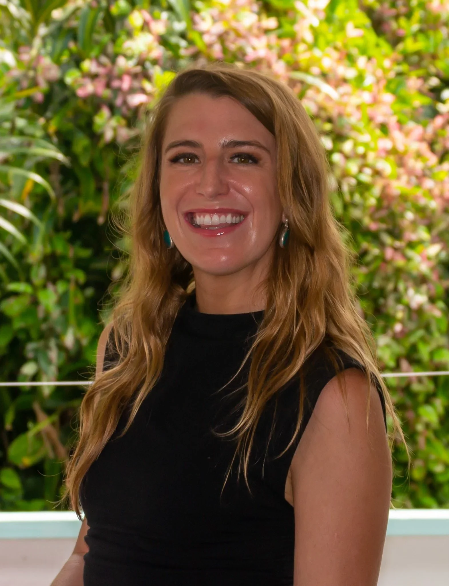 Portrait of a woman with long wavy hair smiling, standing outdoors with pink flowers and green foliage in the background.