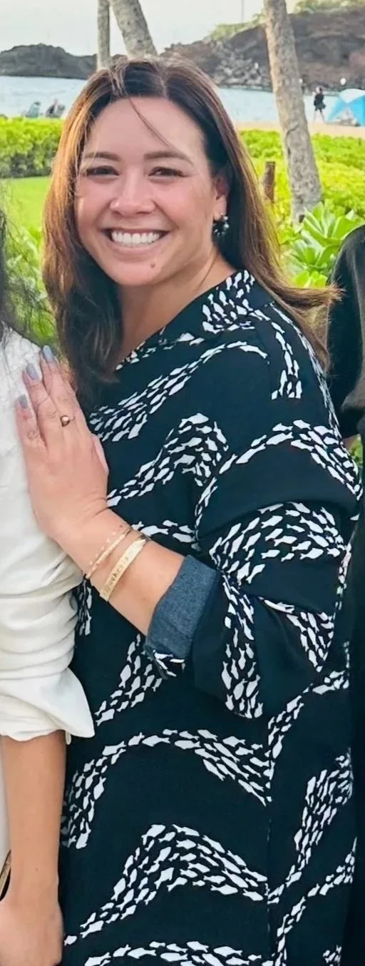 A smiling woman with shoulder-length brown hair, wearing earrings and a black and white patterned dress, standing outdoors near grass, trees, and water.