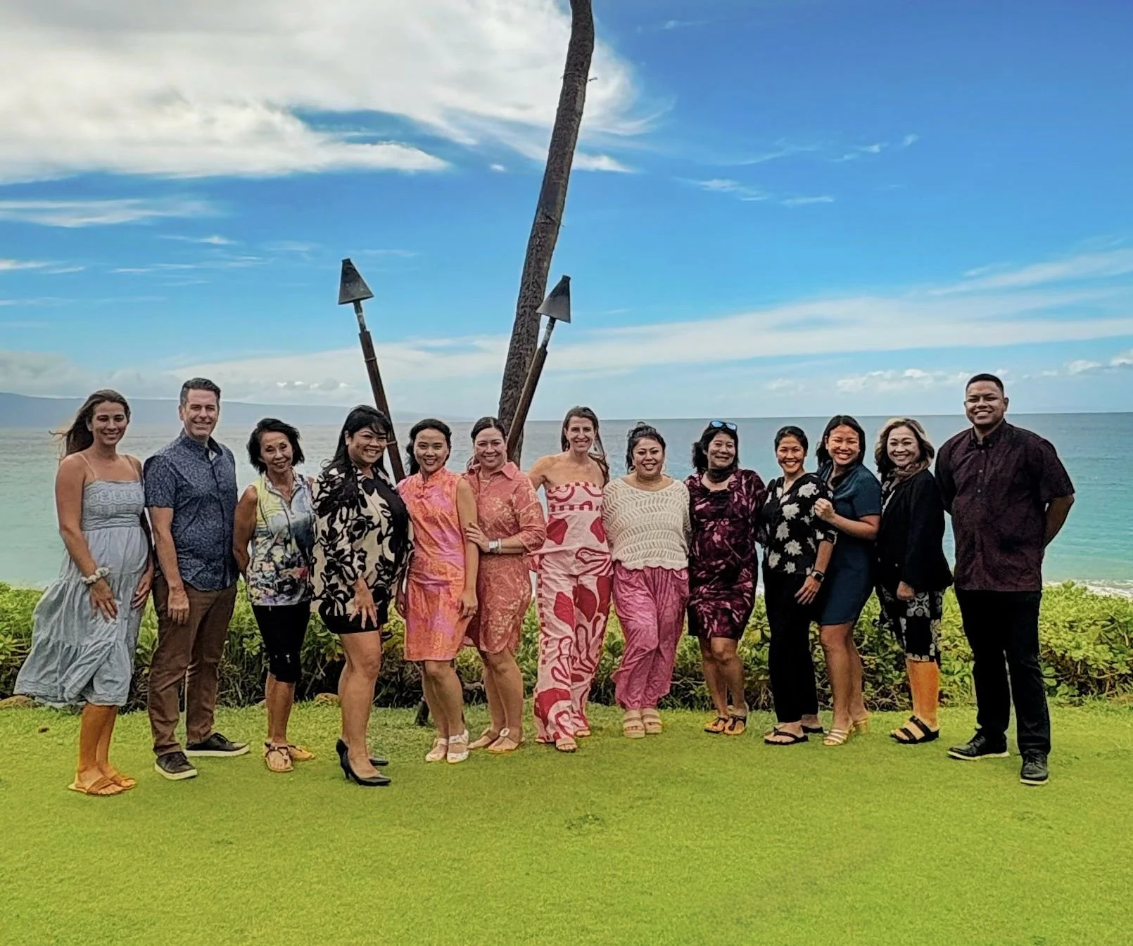 Group of fifteen people standing on grass with ocean and sky in the background, posing for a photo, several holding hands or with arms around one another, some wearing casual and some traditional tropical clothing, a palm tree with two large tiki torches behind them.