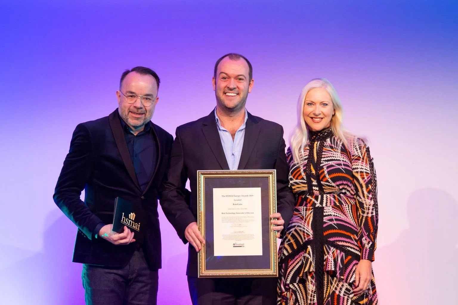 Three people stand together on stage, holding awards and a framed certificate, smiling at the camera. The background is purple.