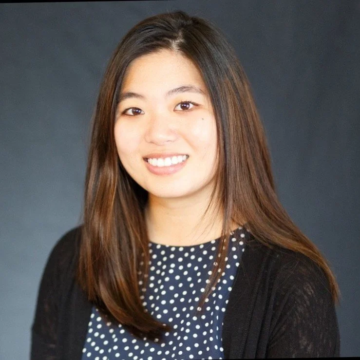A young woman with long brown hair, wearing a navy polka dot top and black cardigan, smiling against a dark background.