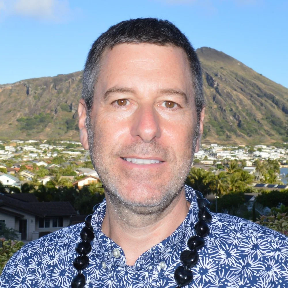A man with short dark hair, light skin, and brown eyes, smiling in front of a mountain and a town with houses and trees, wearing a patterned shirt and a black beaded necklace.