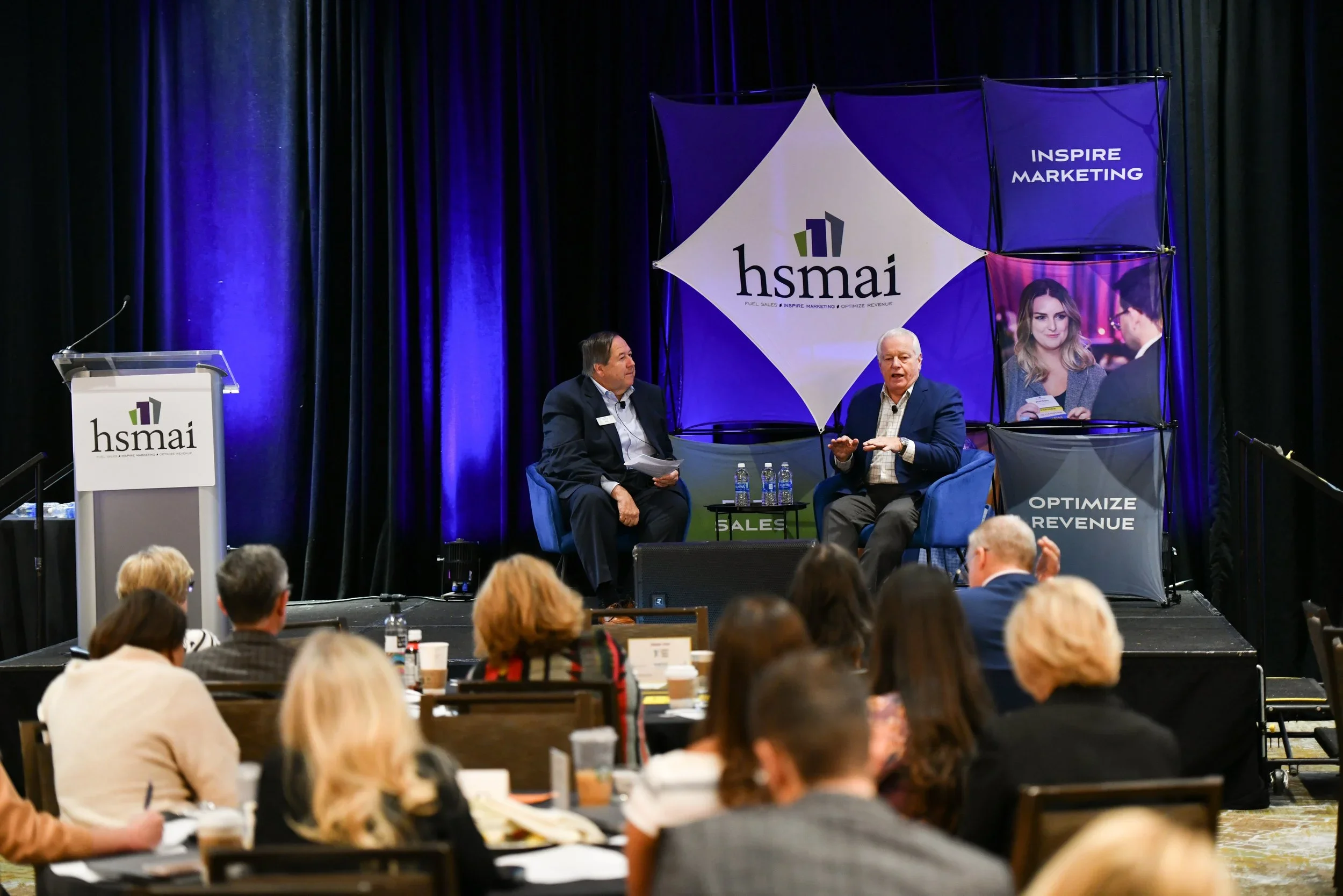 Two men on stage engaged in a discussion at the hsmai conference, with an audience seated in front of them, and a large backdrop displaying the hsmai logo and marketing themes.