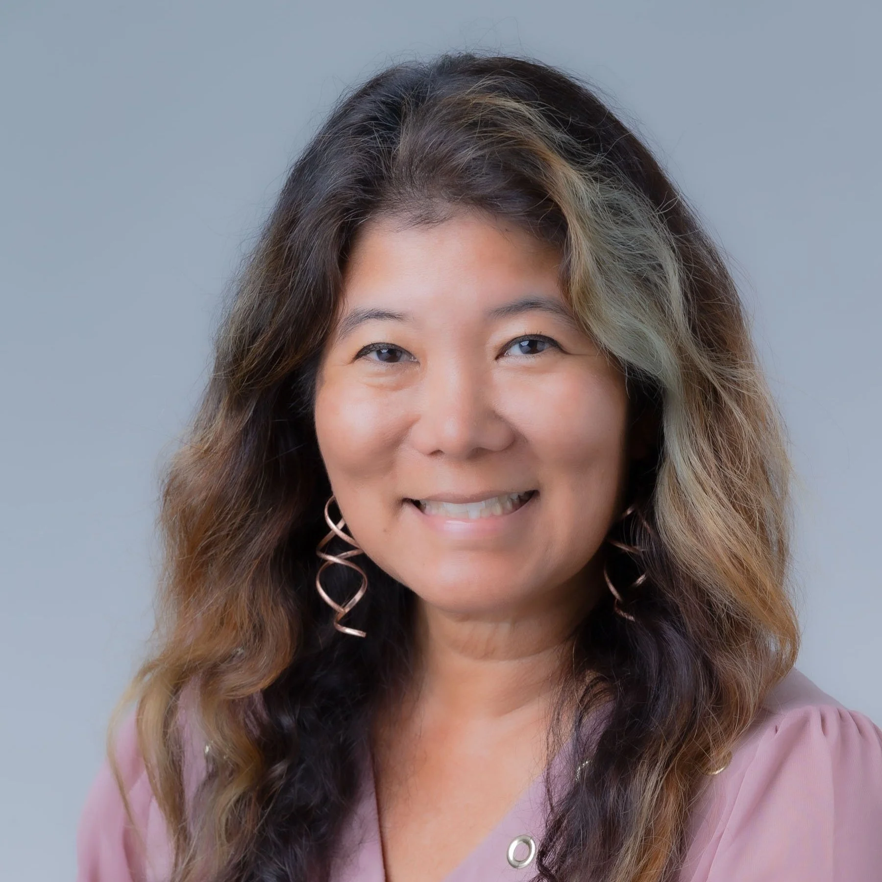 Portrait of a woman with wavy hair, smiling, wearing pink top and spiral earrings against a neutral background.