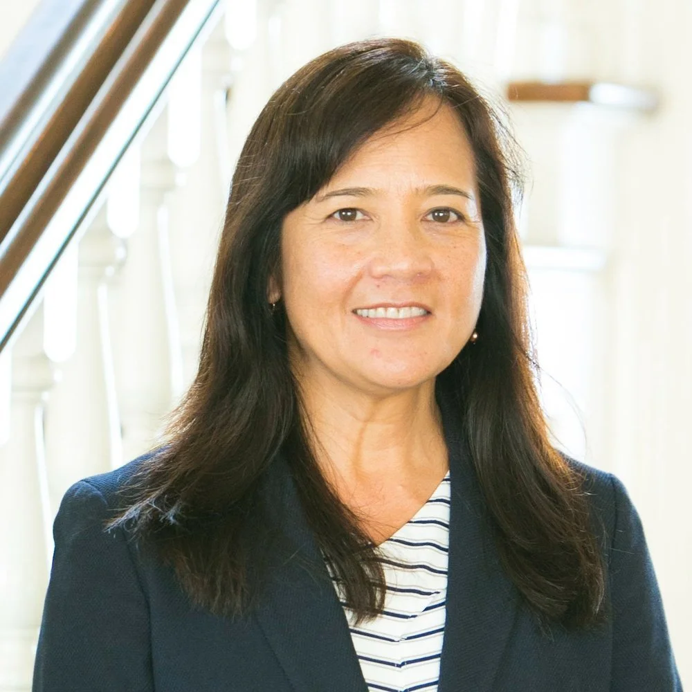 A woman with long dark hair smiling indoors near a staircase, wearing a dark blazer over a striped shirt.