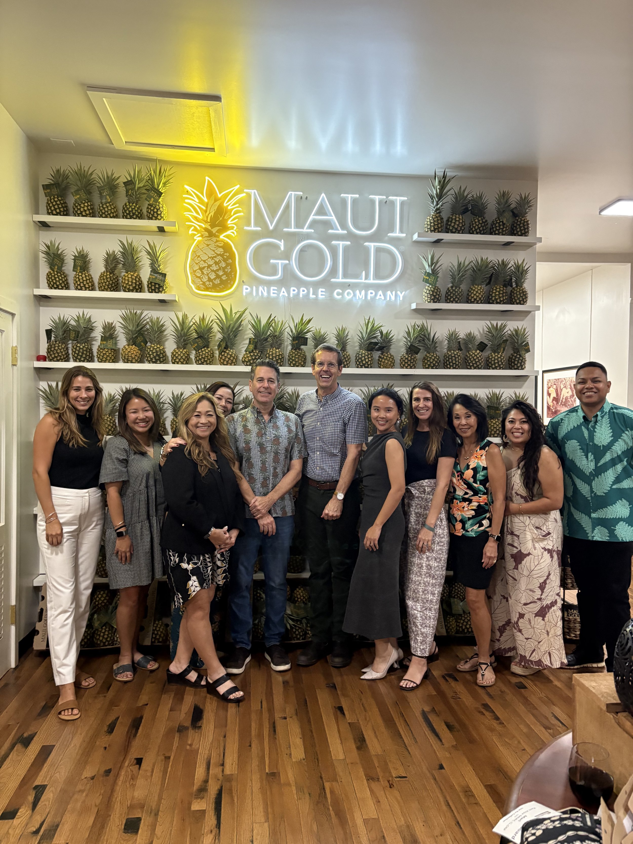 A group of twelve people standing in front of a wall with a neon sign that reads 'Maui Gold Pineapple Company' and multiple pineapples on shelves, inside a store or restaurant.